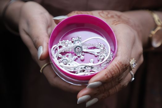 Close-up of intricately designed silver jewelry in a vibrant pink container, held by henna-adorned hands.