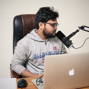 A young man in casual wear recording a podcast at his home office desk with a laptop and microphone.