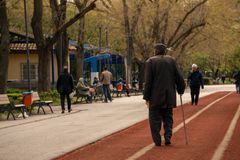 An elderly man with a cane strolls down a serene park path lined with benches and trees.