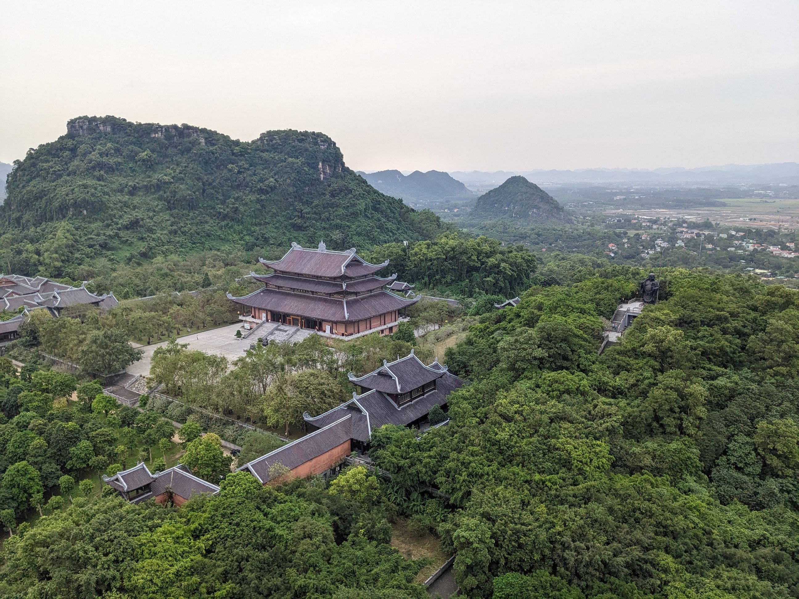 Aerial shot of an Asian temple surrounded by lush greenery and distant mountains, offering a serene landscape.