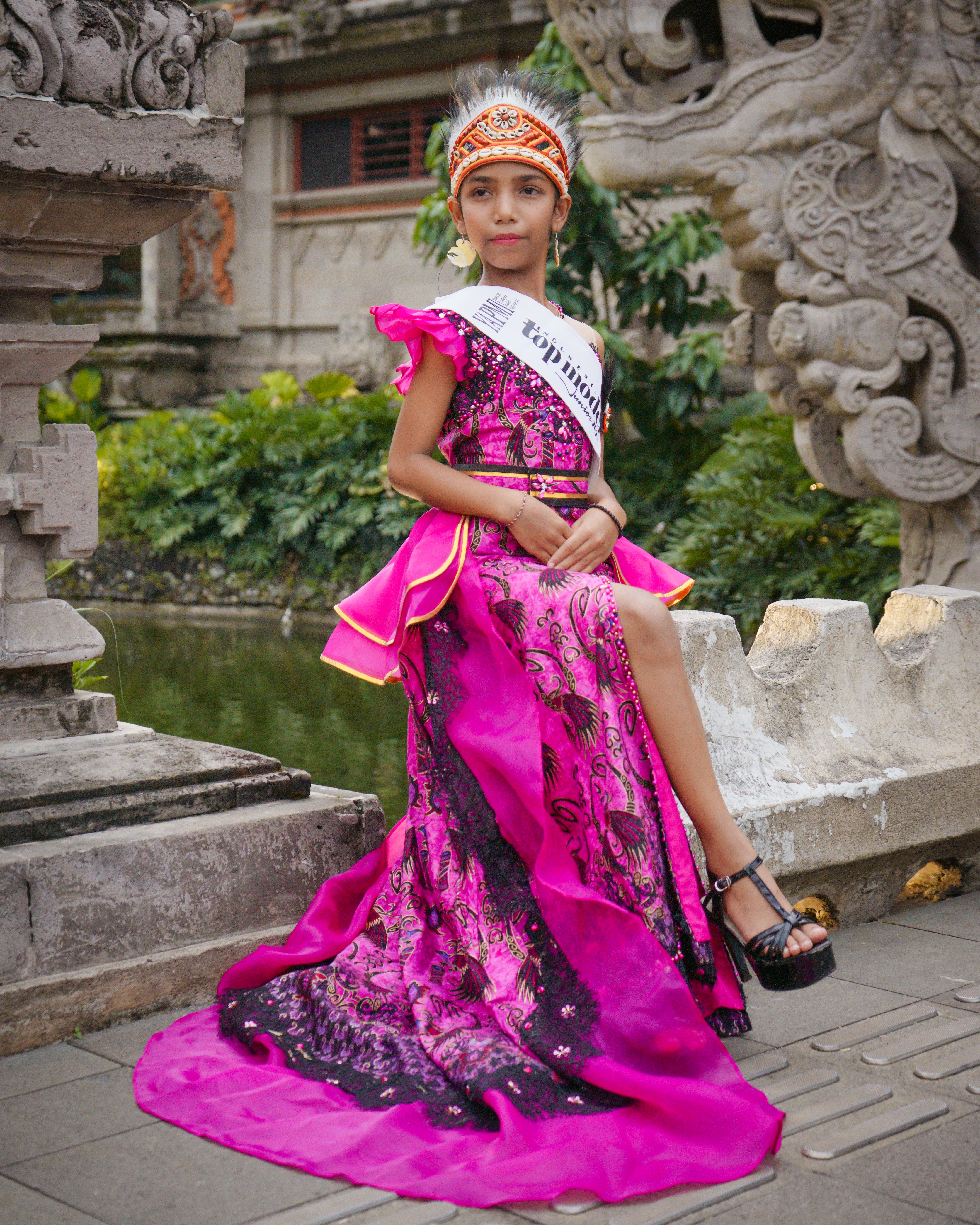 A young girl dressed in a vibrant pink dress poses confidently at a Balinese temple, exuding elegance and culture.