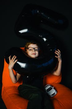 Child with glasses holding number balloon on orange chair, happy celebration.