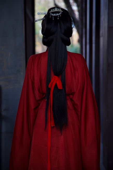 A person in rich red traditional Asian attire poses with intricate hairstyle.