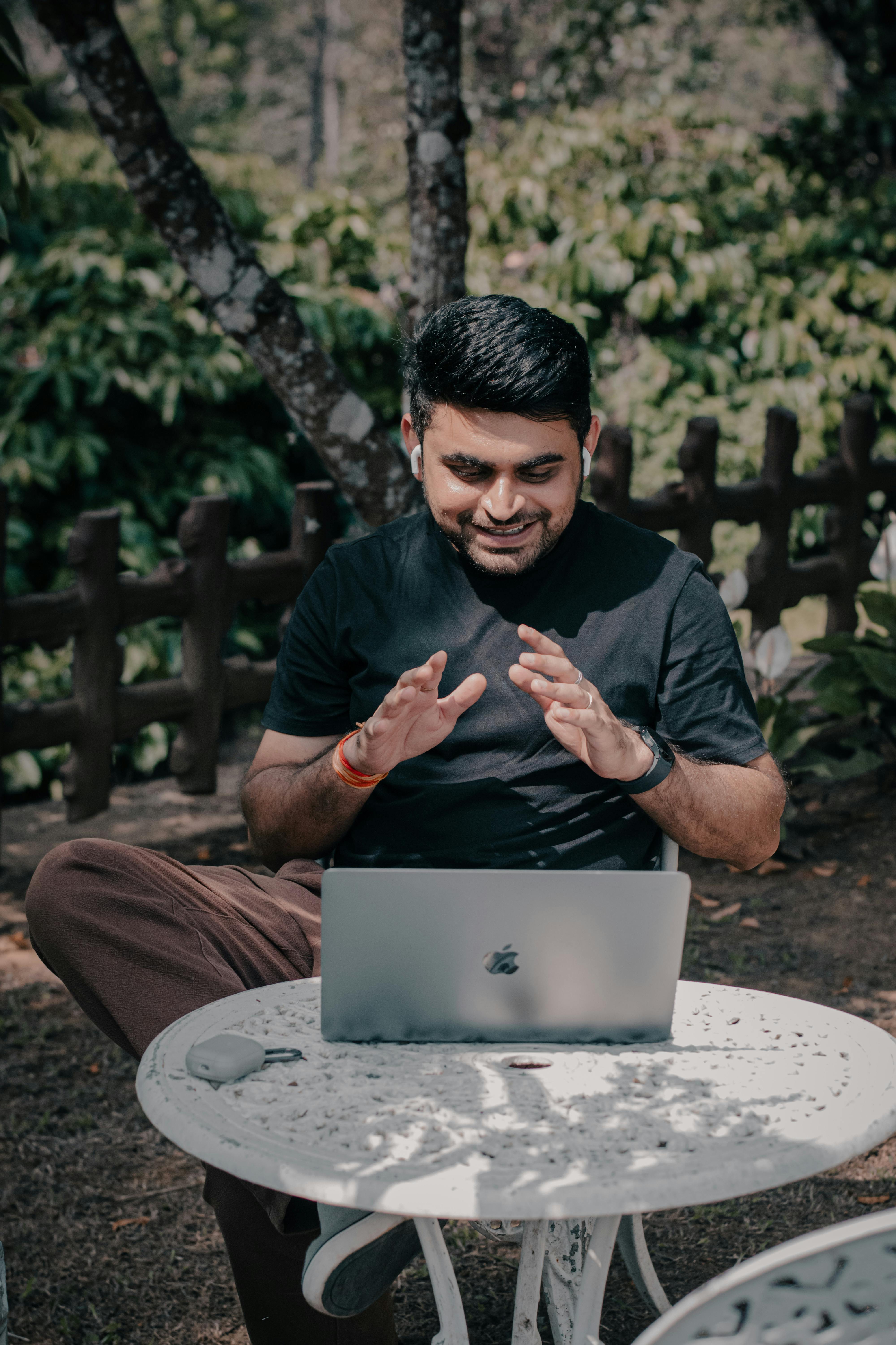 grátis Homem sorridente trabalhando em um laptop em um jardim ao ar livre. Foto profissional