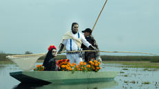 Three people in traditional Day of the Dead attire on a boat with marigold flowers, set in calm waters.