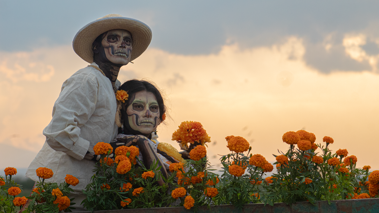 Menschen mit Zuckerschädel-Gesichtsbemalung umgeben von leuchtend orangen Cempasúchil-Ringelblumen beim Día de Muertos in Mexiko