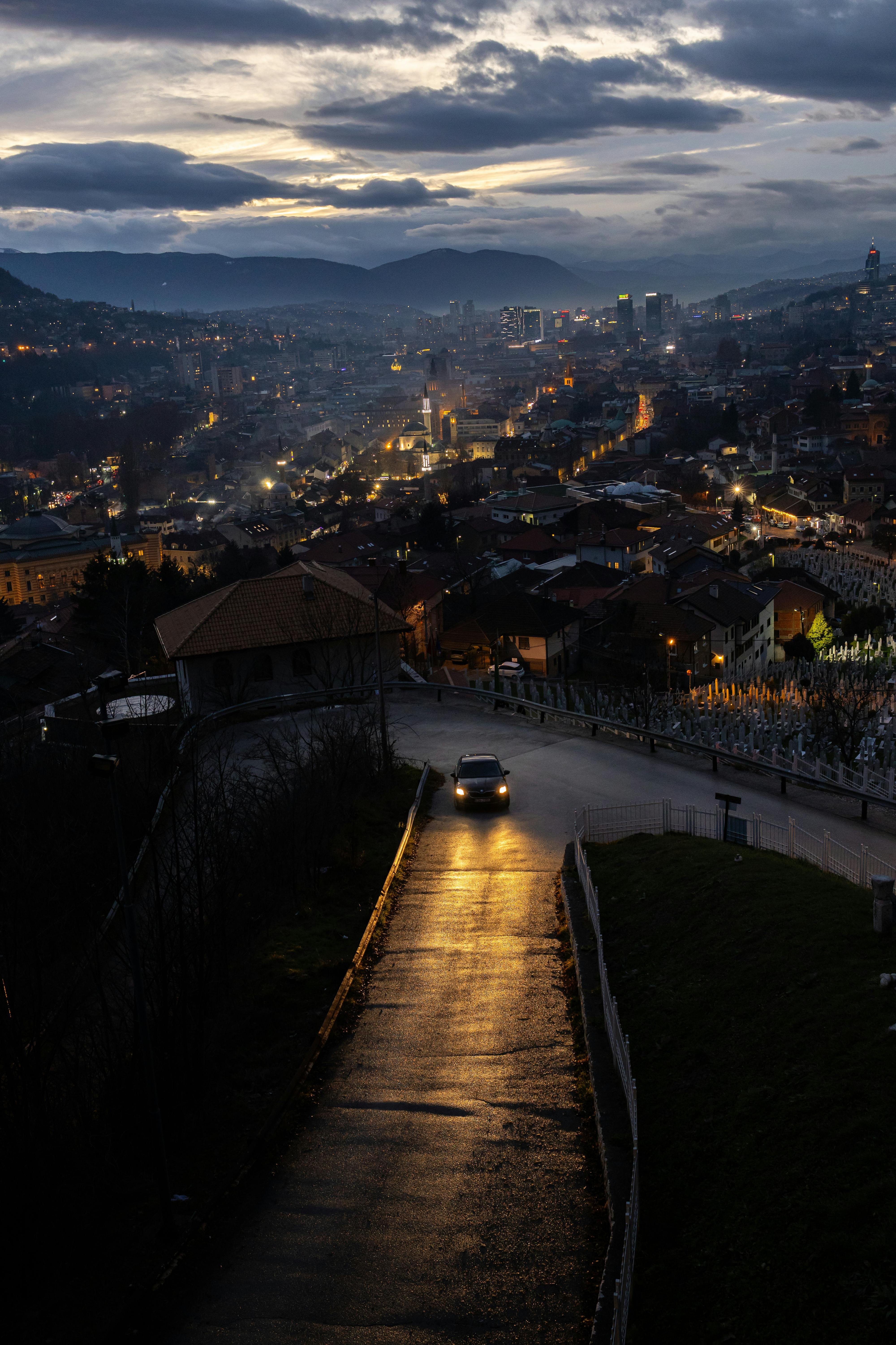 A view over a Bosnian city at dusk with glowing streetlights and a car on a wet road.