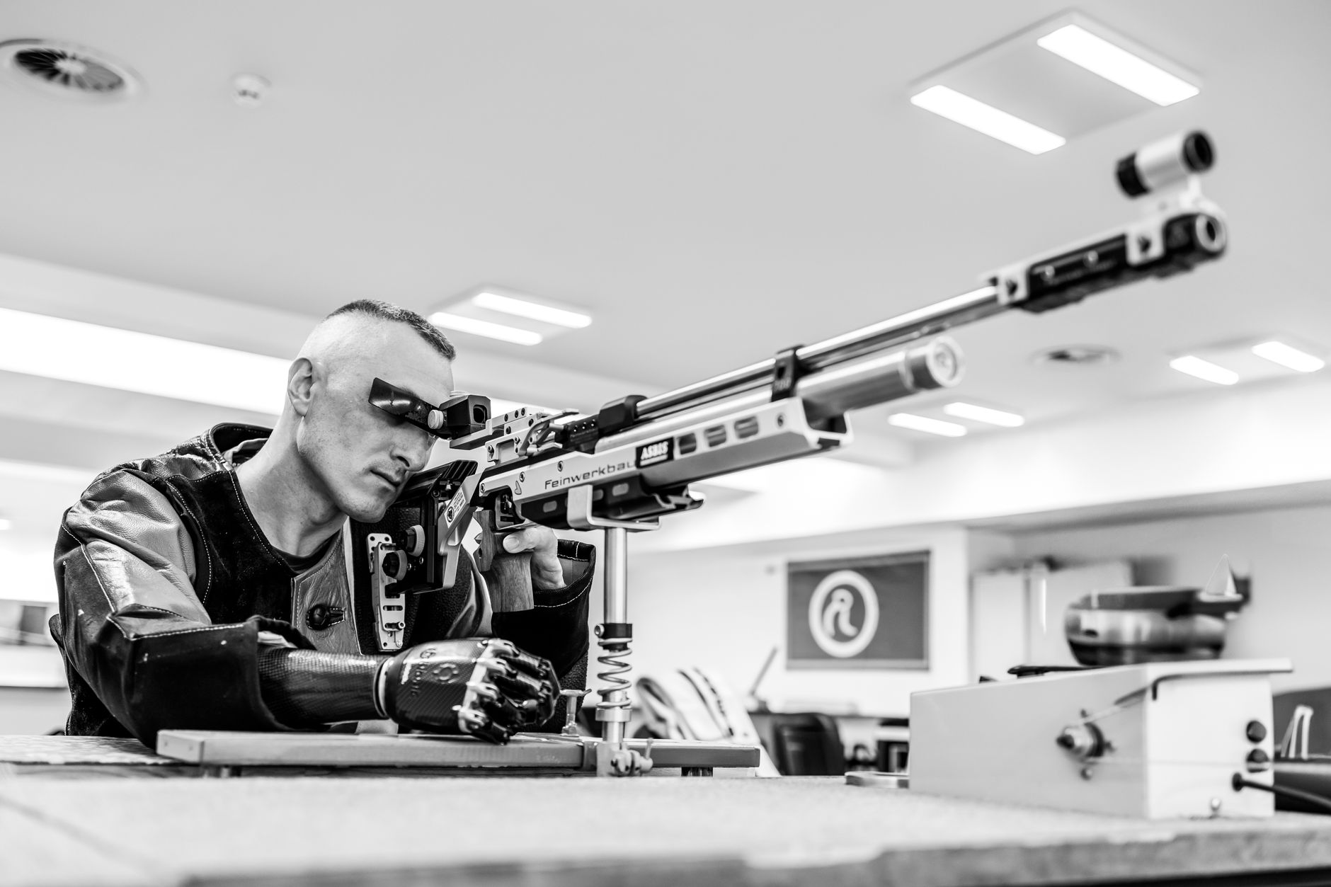 Black and white photo of an air rifle shooter focusing with precision indoors. Competitive sports setting.