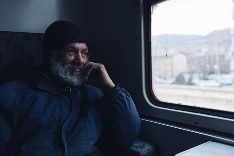A man in a warm jacket and beanie sits in a train, gazing thoughtfully out the window.