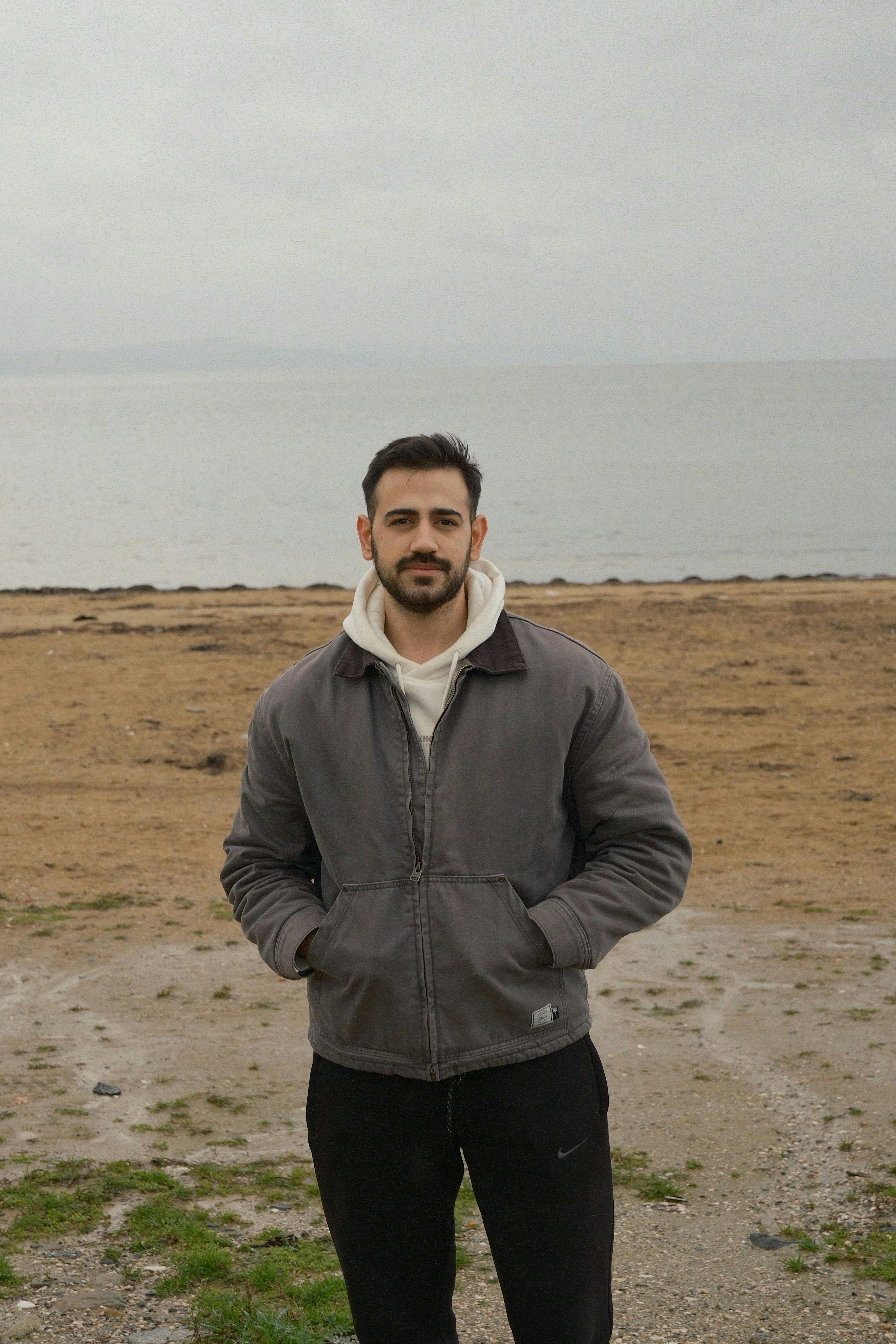 Man in a jacket stands on a quiet beach, cloudy sky overhead.