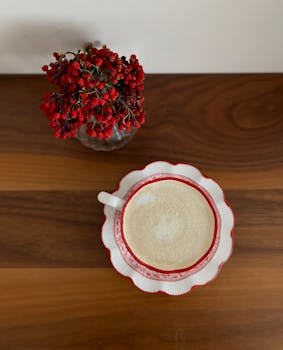 A cappuccino in a red-rimmed cup sits beside a vase of red berries on a wooden surface, exuding tranquility.