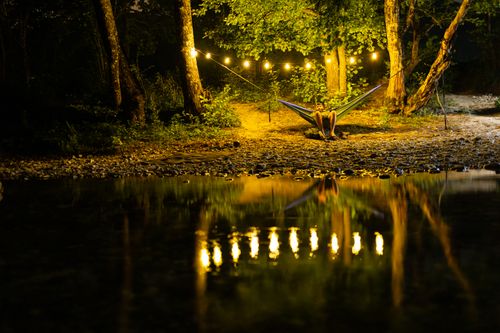 Free Person relaxing in a hammock at night with string lights, reflecting on a tranquil lakeside setting. Stock Photo