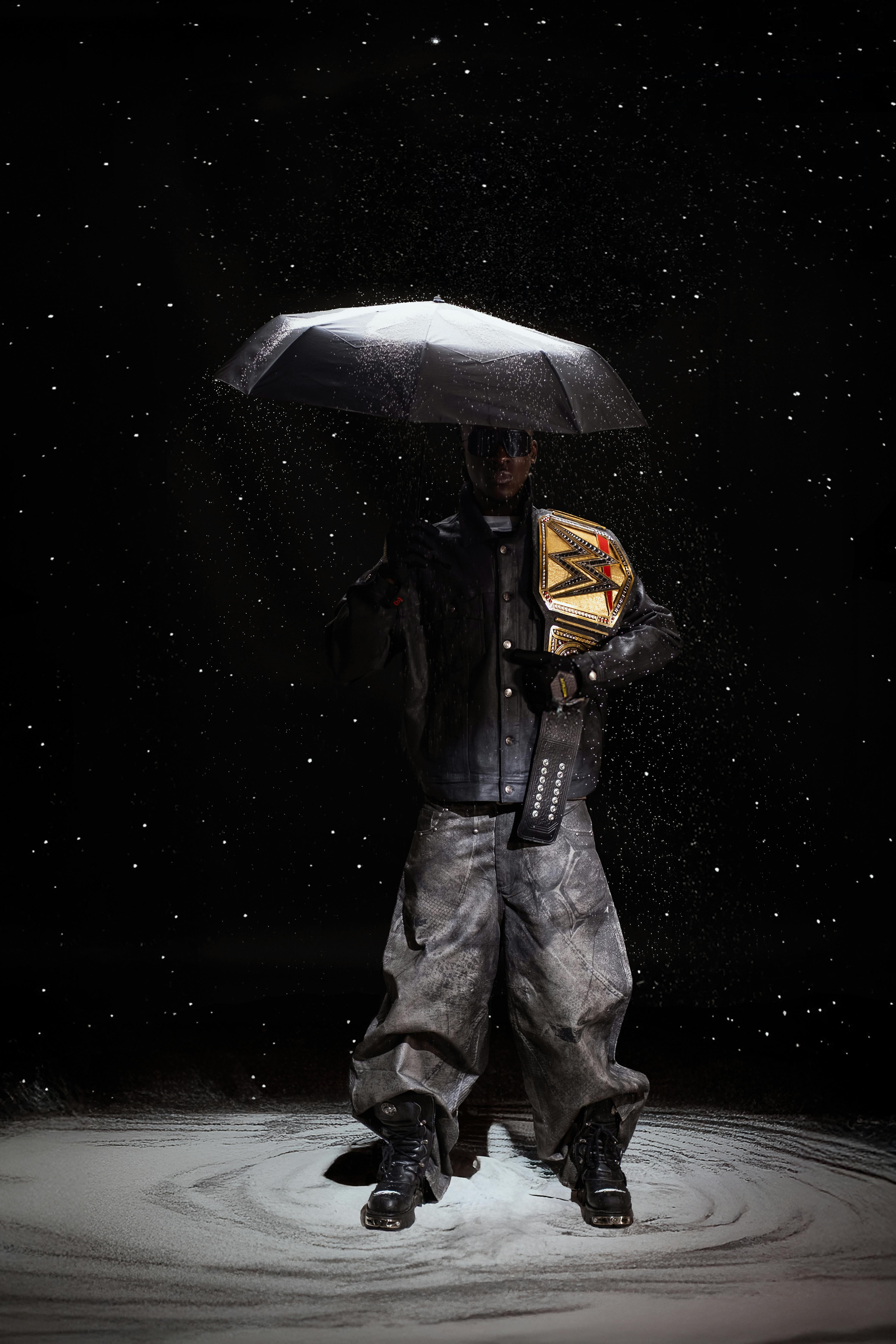 A champion in leather stands with an umbrella and belt under falling snow in dramatic lighting.
