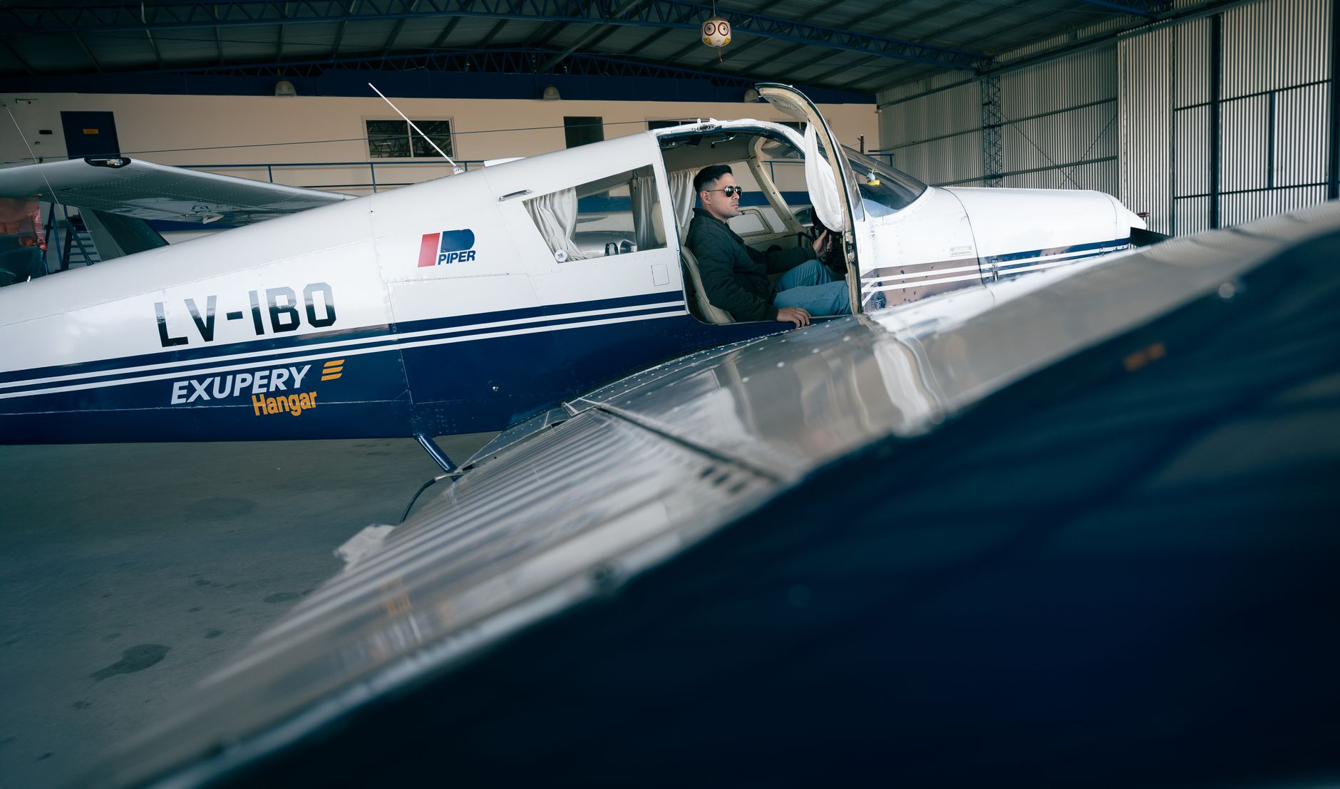 Pilot in a Piper aircraft with hangar setting, ready for takeoff.