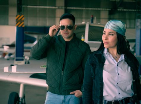 Two individuals in a hangar, sunglasses and casual attire, near aircraft.