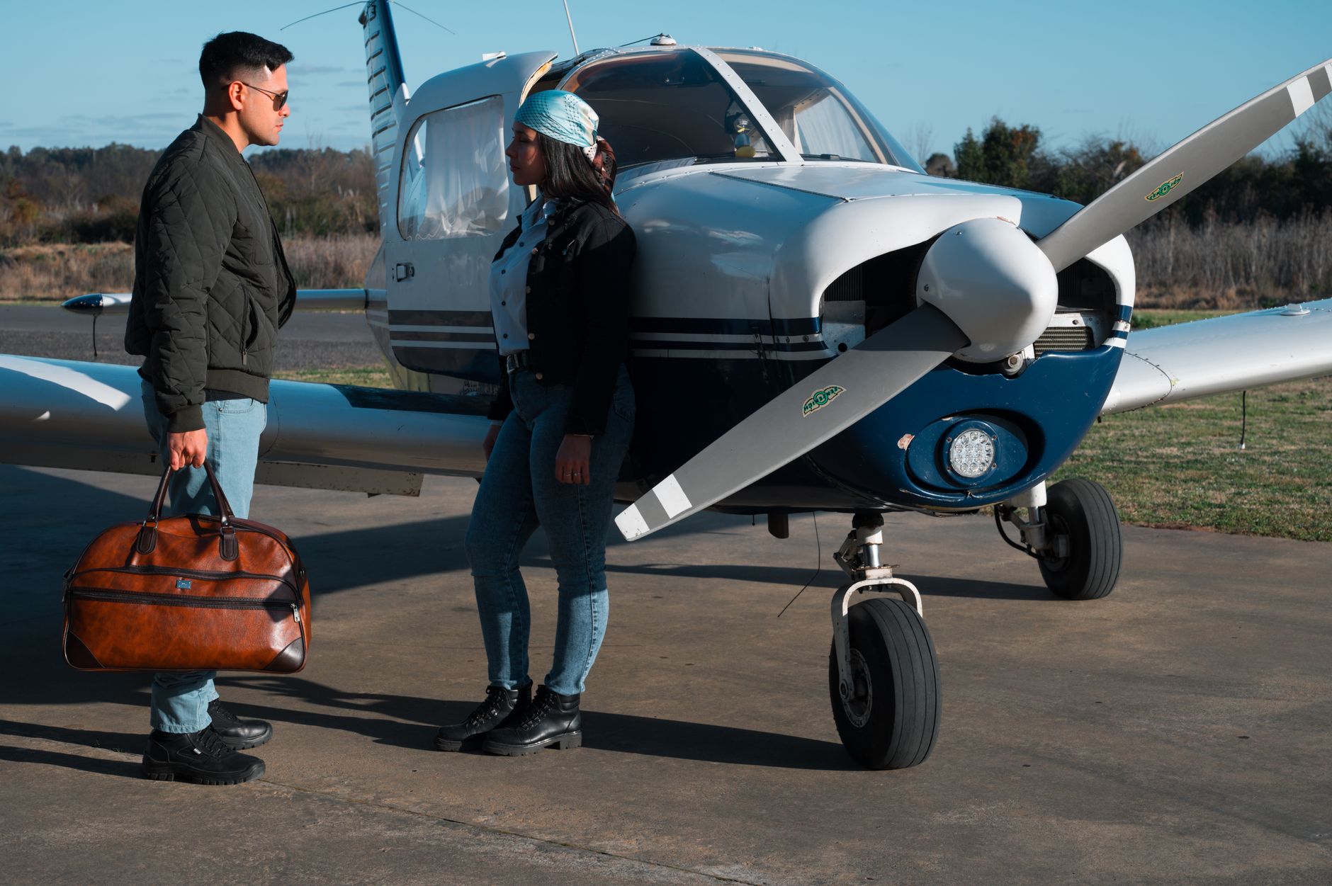 Young couple standing by a private airplane, conversing and holding a bag, at a sunny airfield.