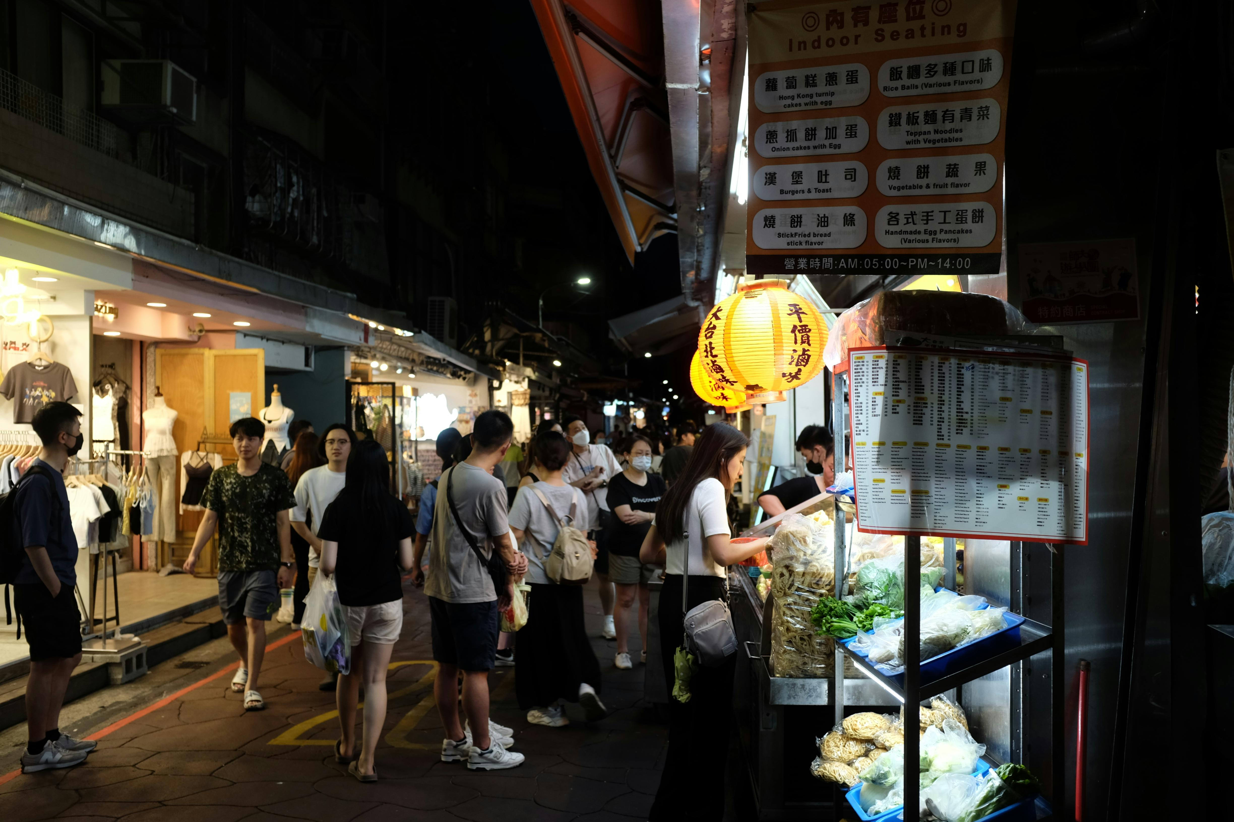 Crowded night market scene with people shopping and dining under bright lanterns.