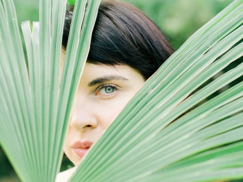 Free A woman peeks through vibrant green palm leaves, set against a natural outdoor backdrop. Stock Photo