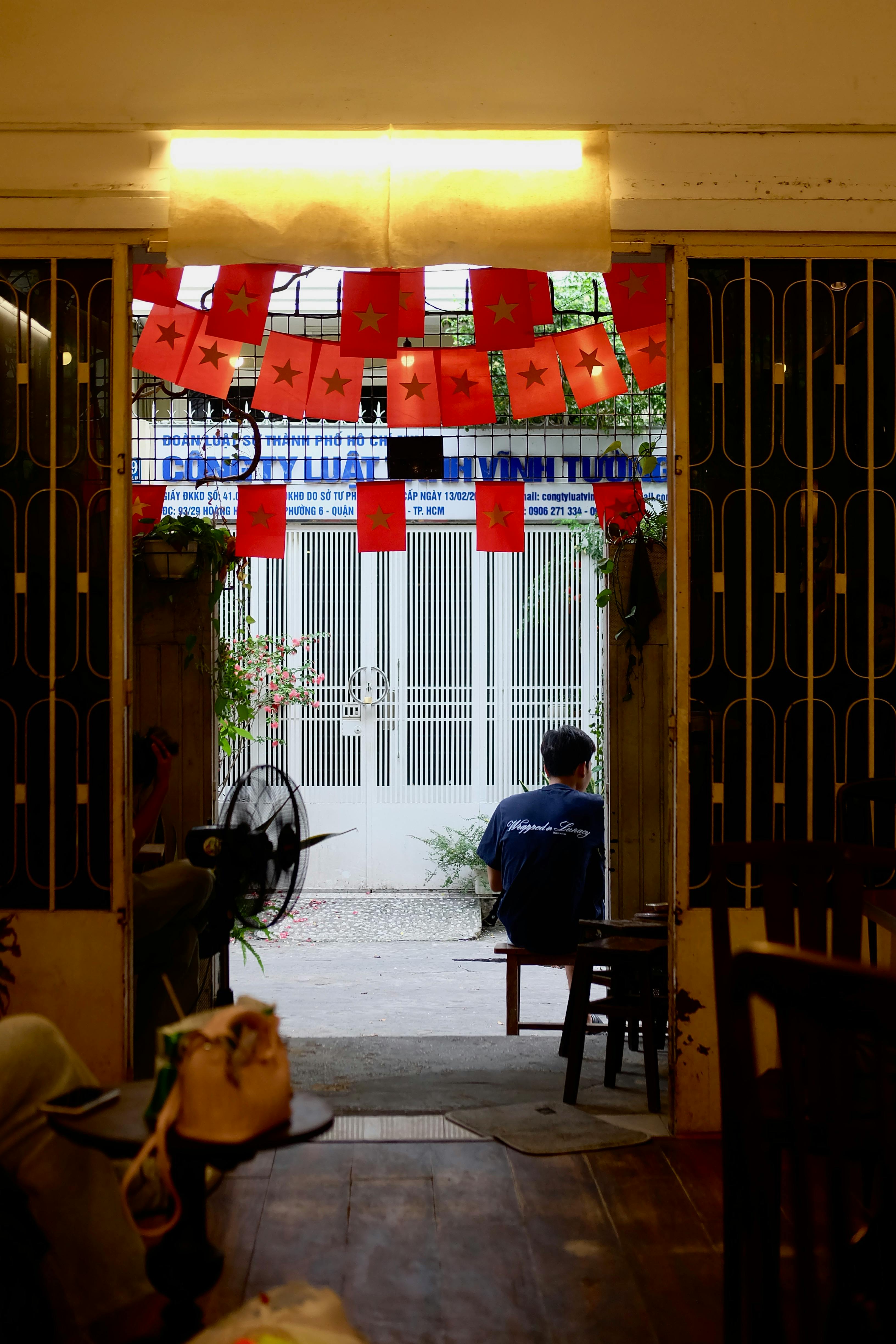 Gratuit O vedere liniștită a unei cafenele interioare, cu bannere roșii decorative și o persoană așezată lângă ușa deschisă. Fotografie de stoc