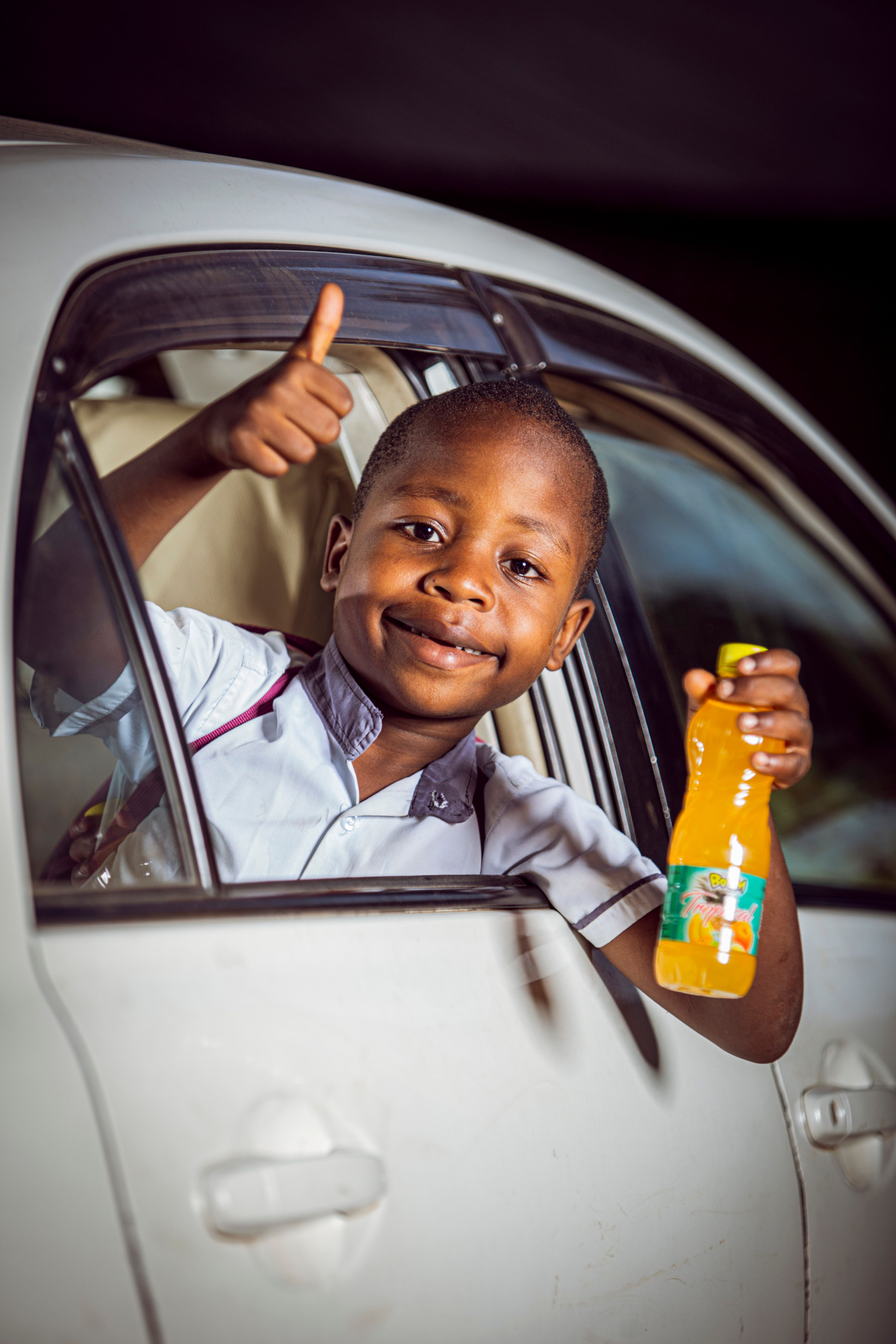 Smiling child gives thumbs up holding orange juice in car window.