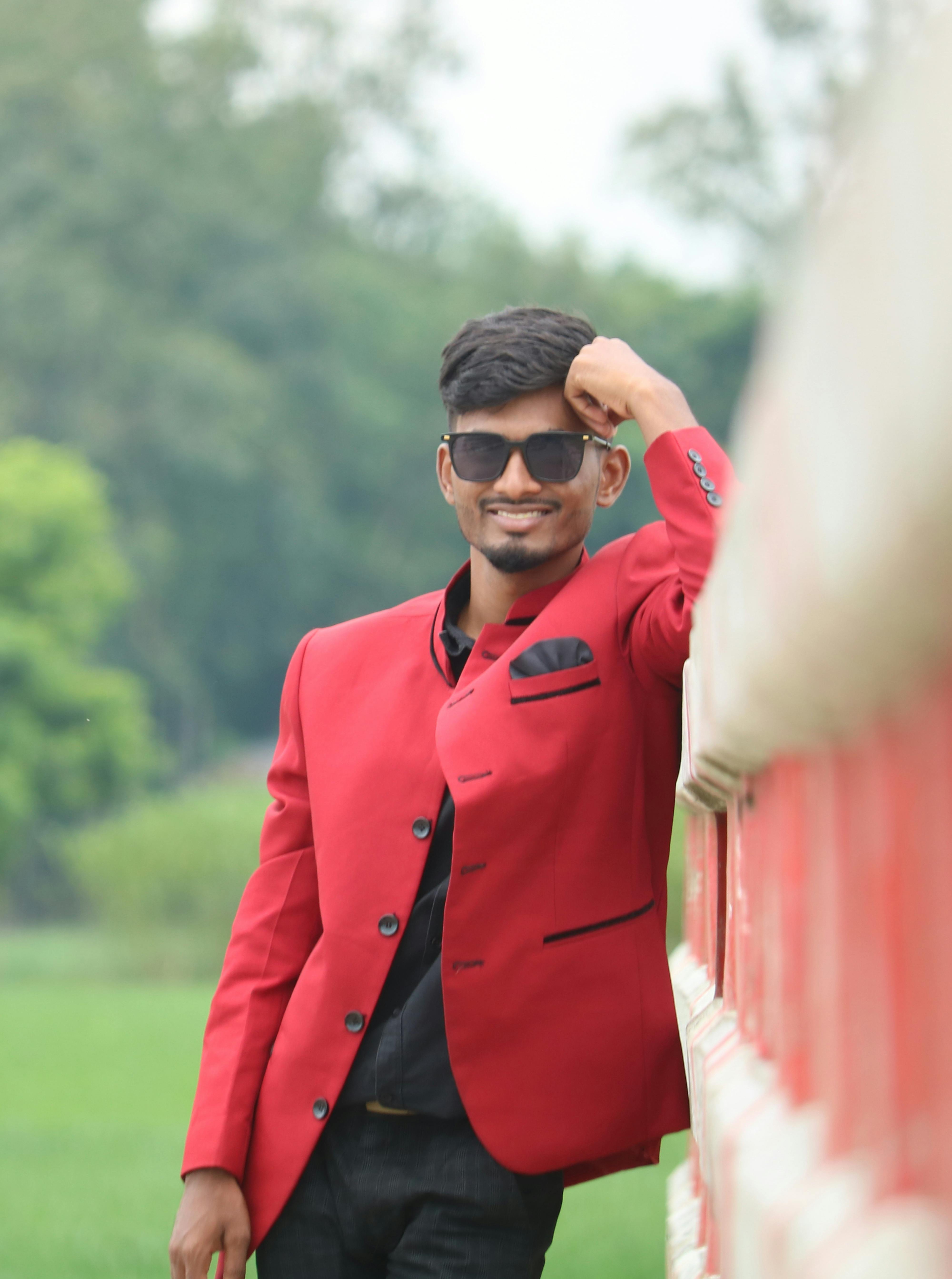 Confident man in red jacket and sunglasses leaning on a fence in a lush green outdoor setting.