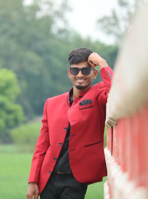 Confident man in red jacket and sunglasses leaning on a fence in a lush green outdoor setting.
