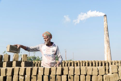 Free An elderly man works at a brick kiln, stacking handmade bricks under a clear blue sky. Stock Photo