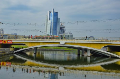 Free A scenic view of a modern city bridge over a reflective river with buildings and people in the background. Stock Photo