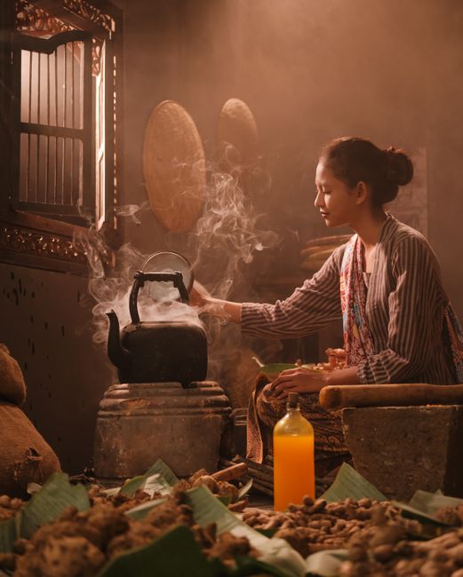 Asian woman in traditional attire preparing herbal tea indoors with steam rising from kettle.