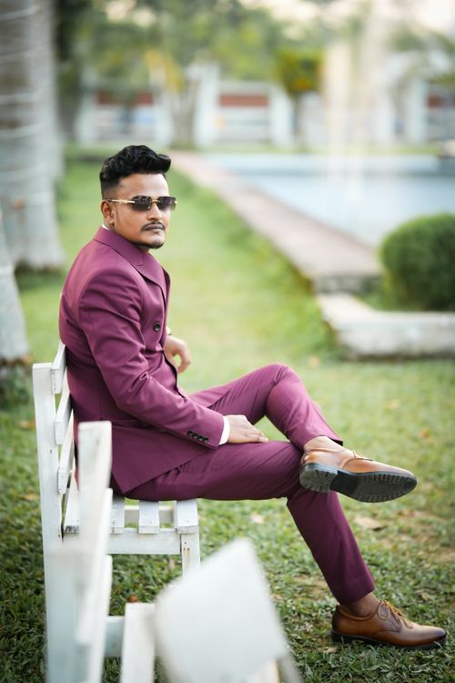 Free Confident man in a maroon suit sitting on a white bench outdoors with trees and grass. Stock Photo