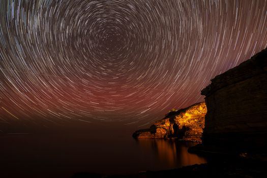 Mesmerizing long-exposure star trails above an illuminated cliffside by the ocean.