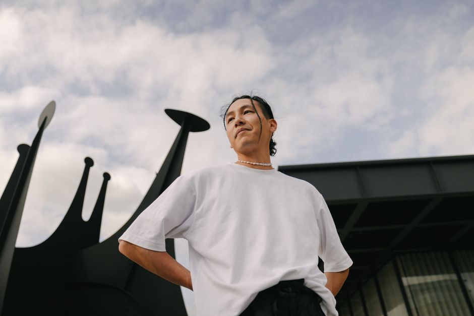 Stylish individual posing against abstract sculpture under cloudy sky.