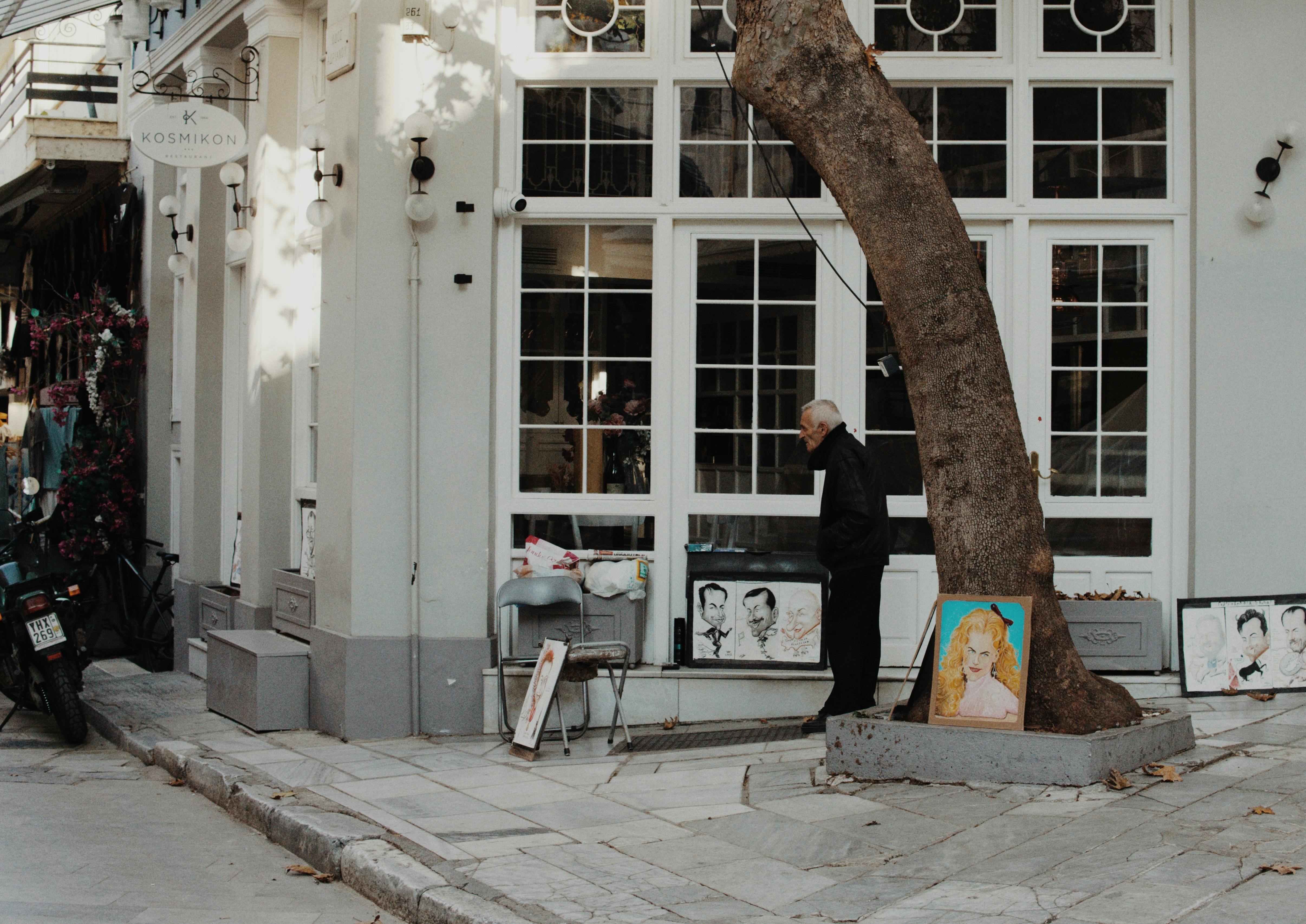 An elderly artist showcases portraits on a city street under a tree.
