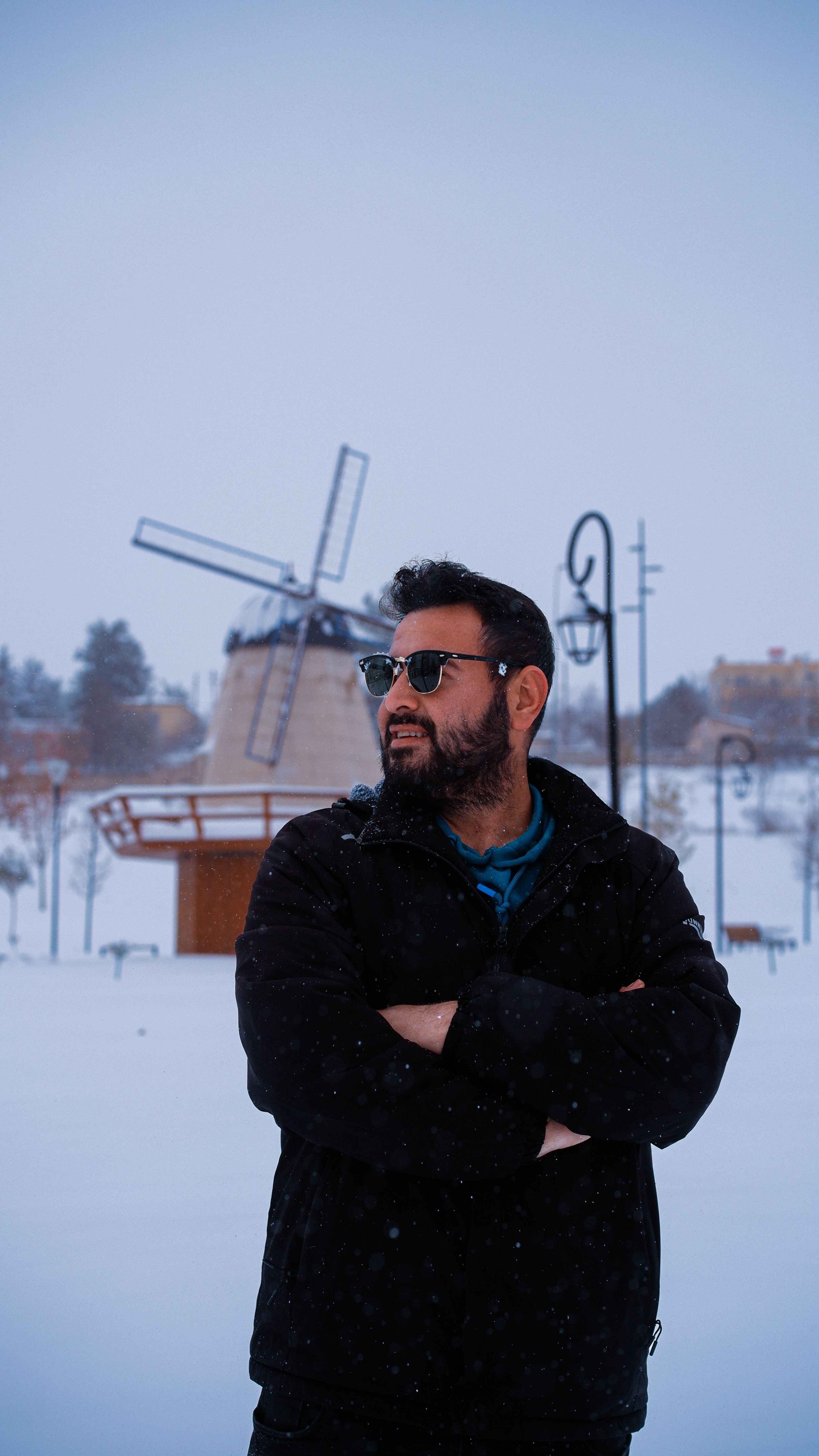 Man in sunglasses standing in snowy landscape with windmill in background.