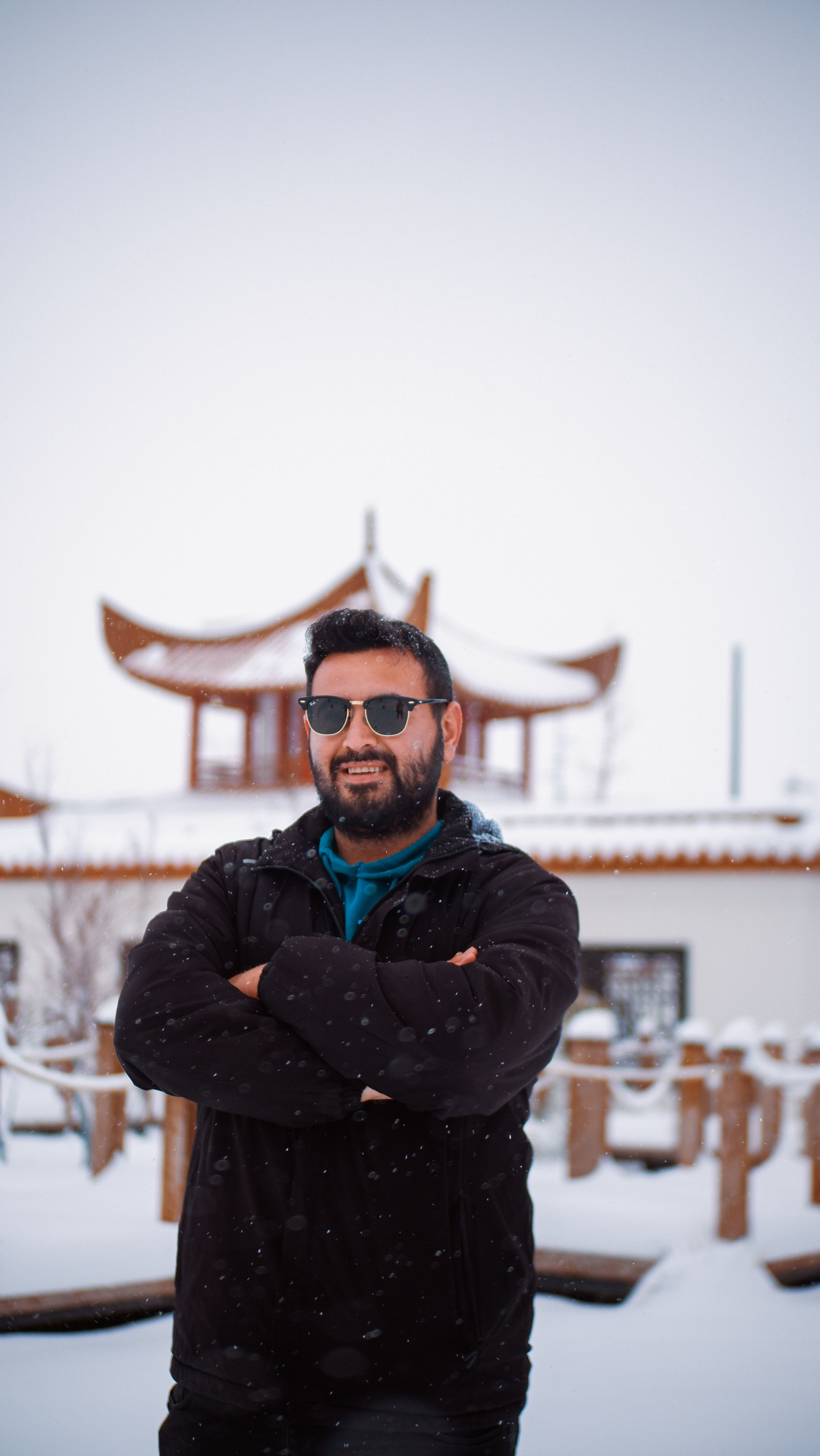 Man in sunglasses and winter coat stands in snow with traditional pagoda behind.