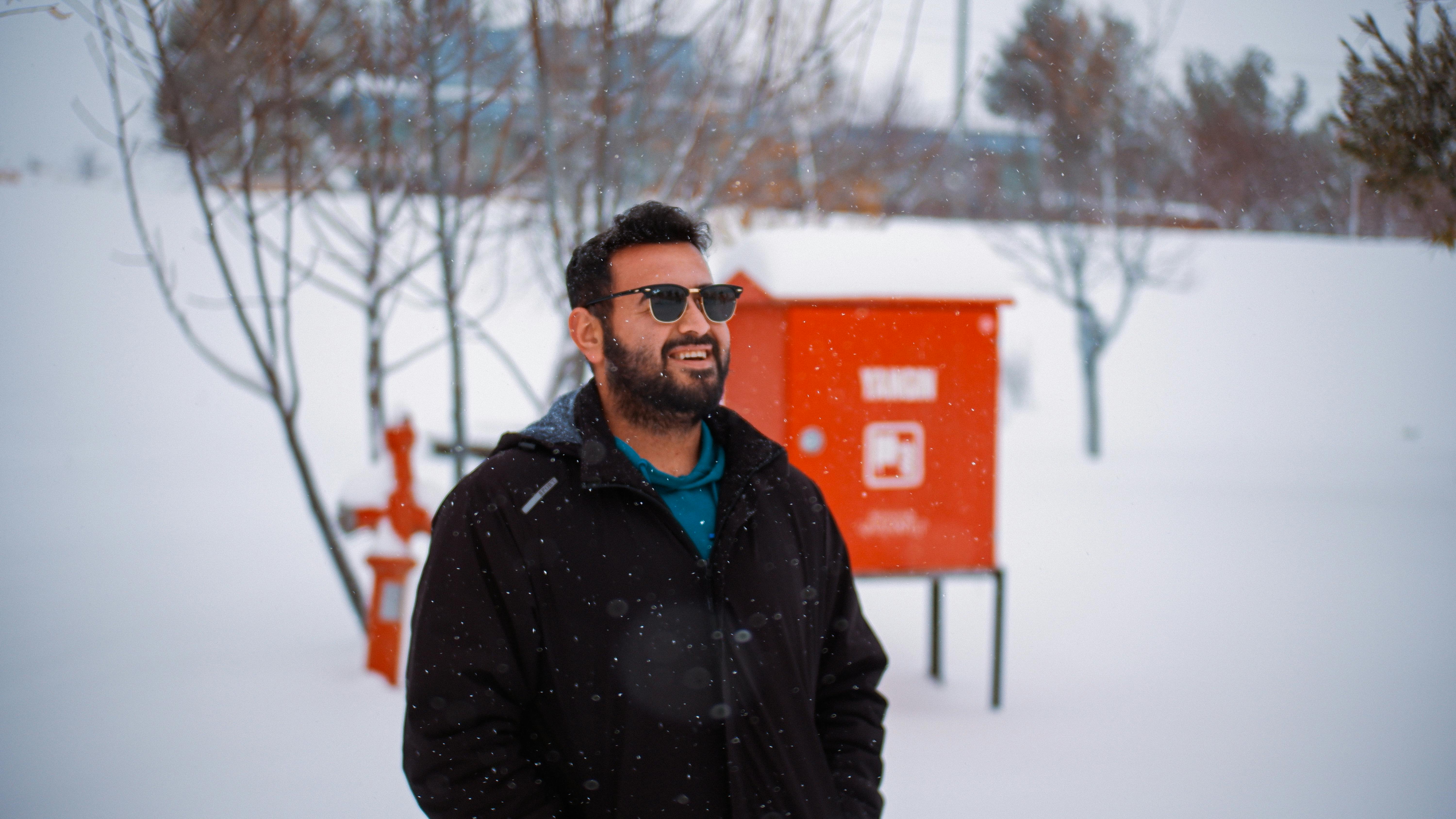 A man in sunglasses and a coat smiles while enjoying a snowy winter day outdoors.