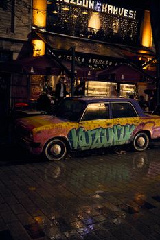 Graffiti-covered car parked near illuminated cafe on a city street at night.