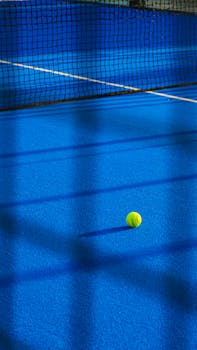 Tennis ball on a vibrant blue court, casting shadows under daylight.