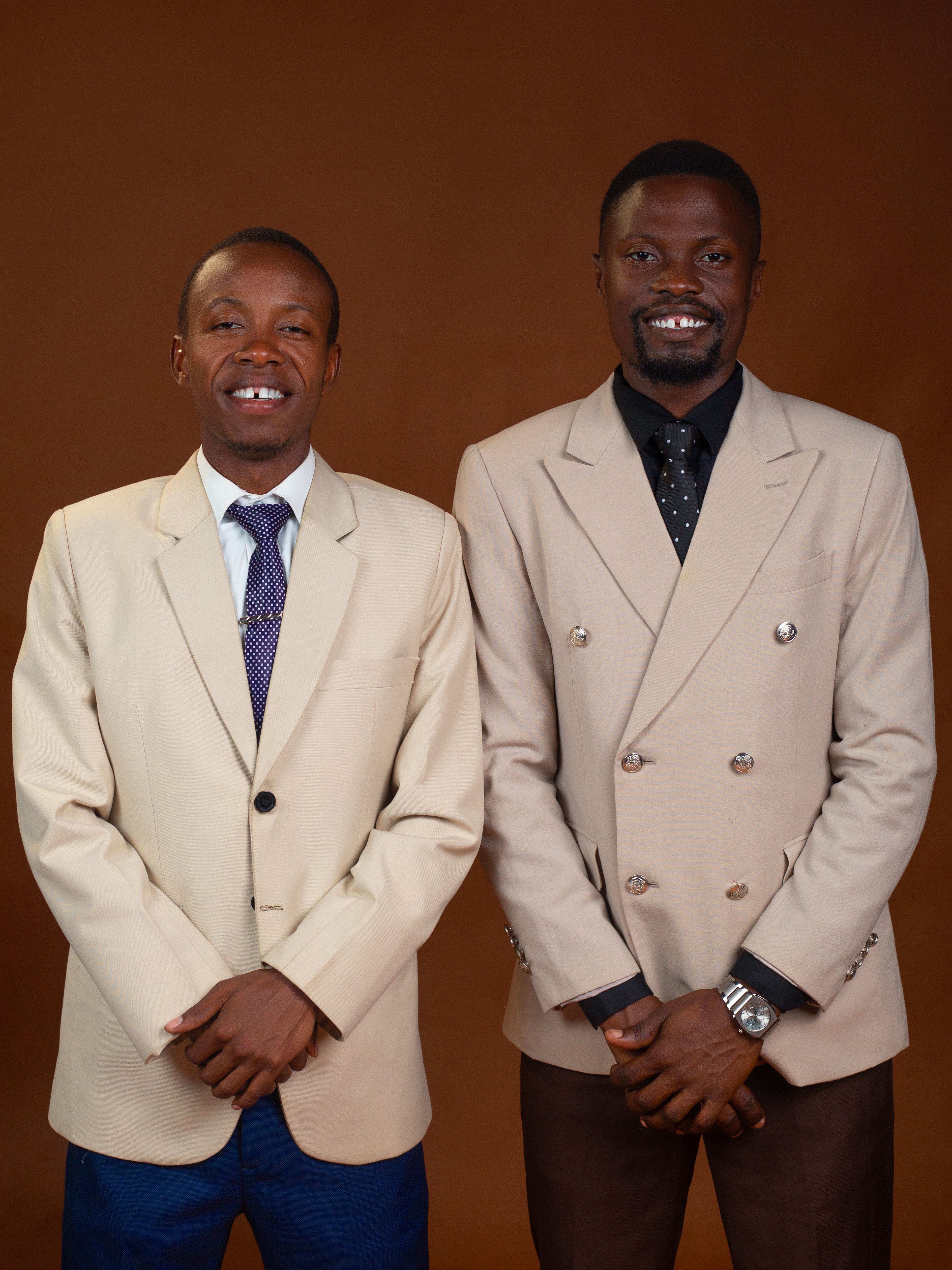 Two men smiling, wearing beige suits in a studio setting against a brown background.