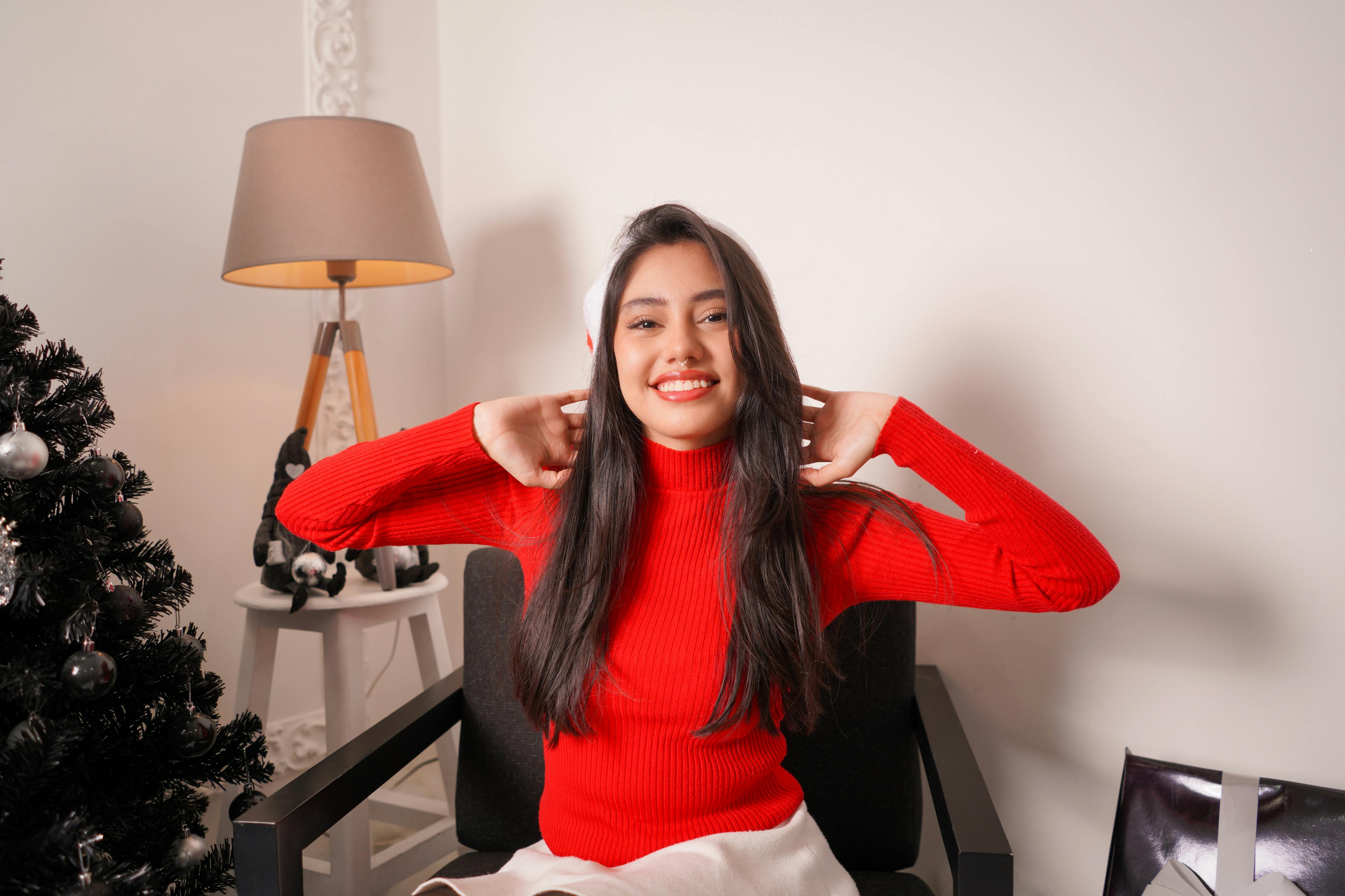 grátis Jovem sorridente de suéter vermelho aproveitando o Natal dentro de casa perto da árvore decorada. Foto profissional