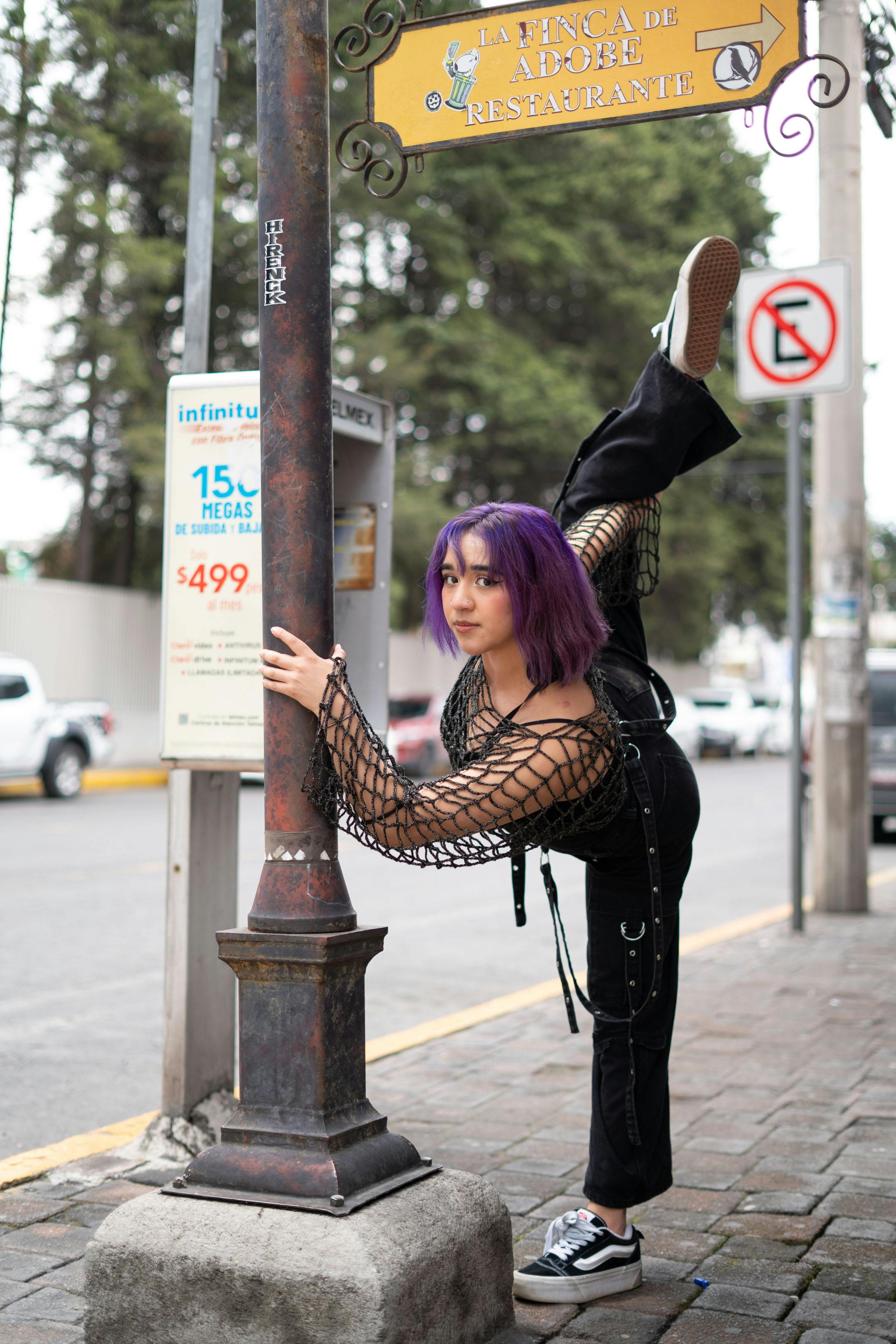 grátis Jovem mulher com roupa estilosa executa uma pose de chute alto enquanto se apoia em um poste de rua em um ambiente urbano. Foto profissional