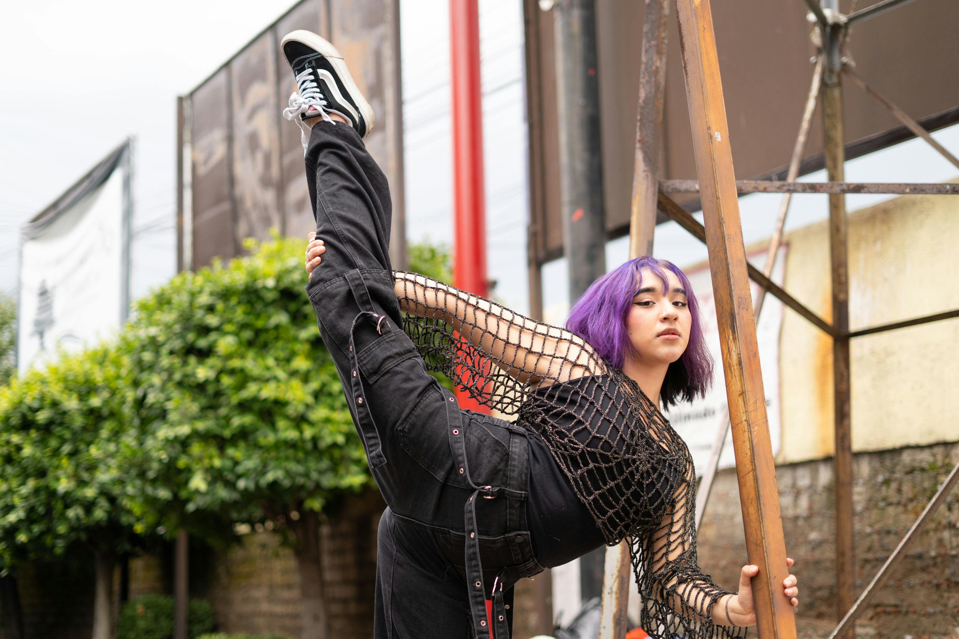 grátis Dançarina de rua confiante, com cabelo roxo, executa um chute alto em um ambiente urbano, demonstrando flexibilidade e estilo. Foto profissional