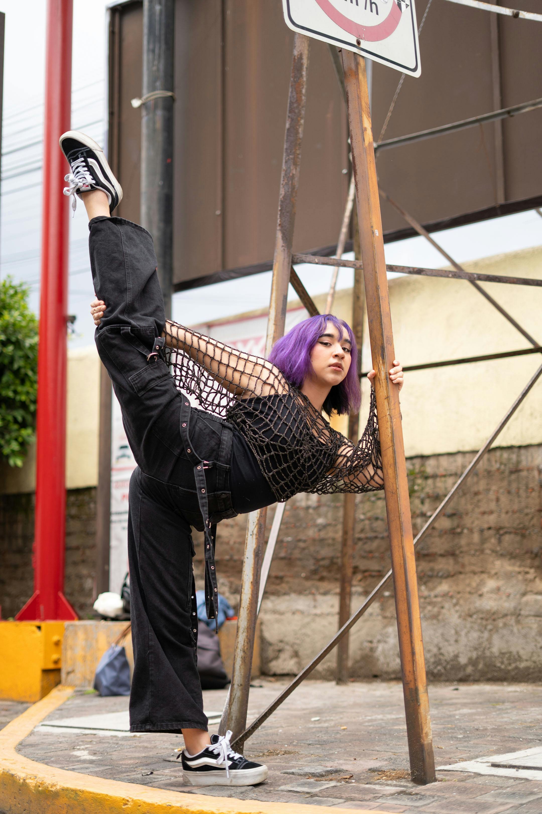 grátis Jovem mulher com vibrantes cabelos roxos demonstrando flexibilidade em um ambiente urbano de rua. Foto profissional