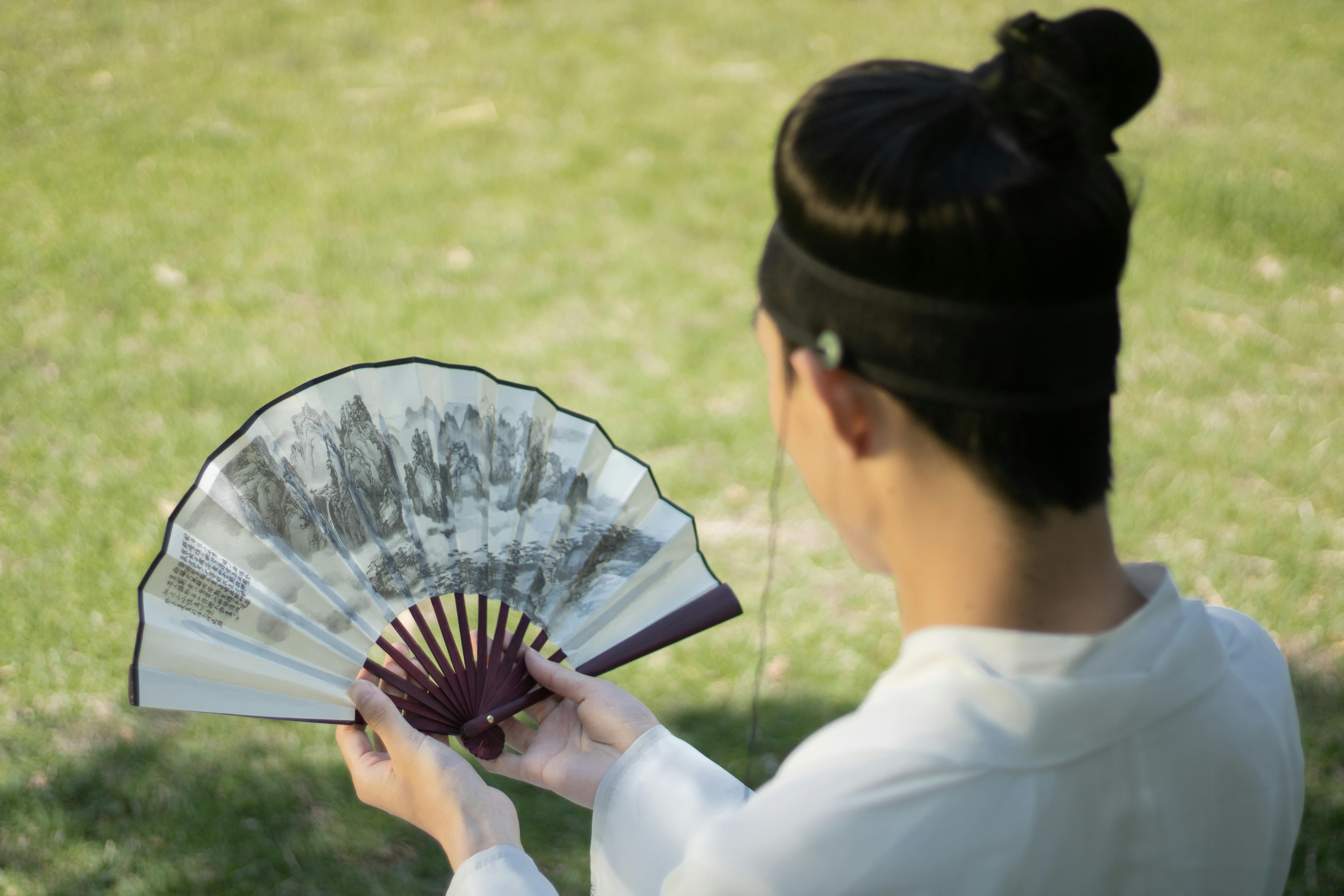 grátis Um homem em trajes tradicionais segurando um leque chinês decorado em um parque. Foto profissional