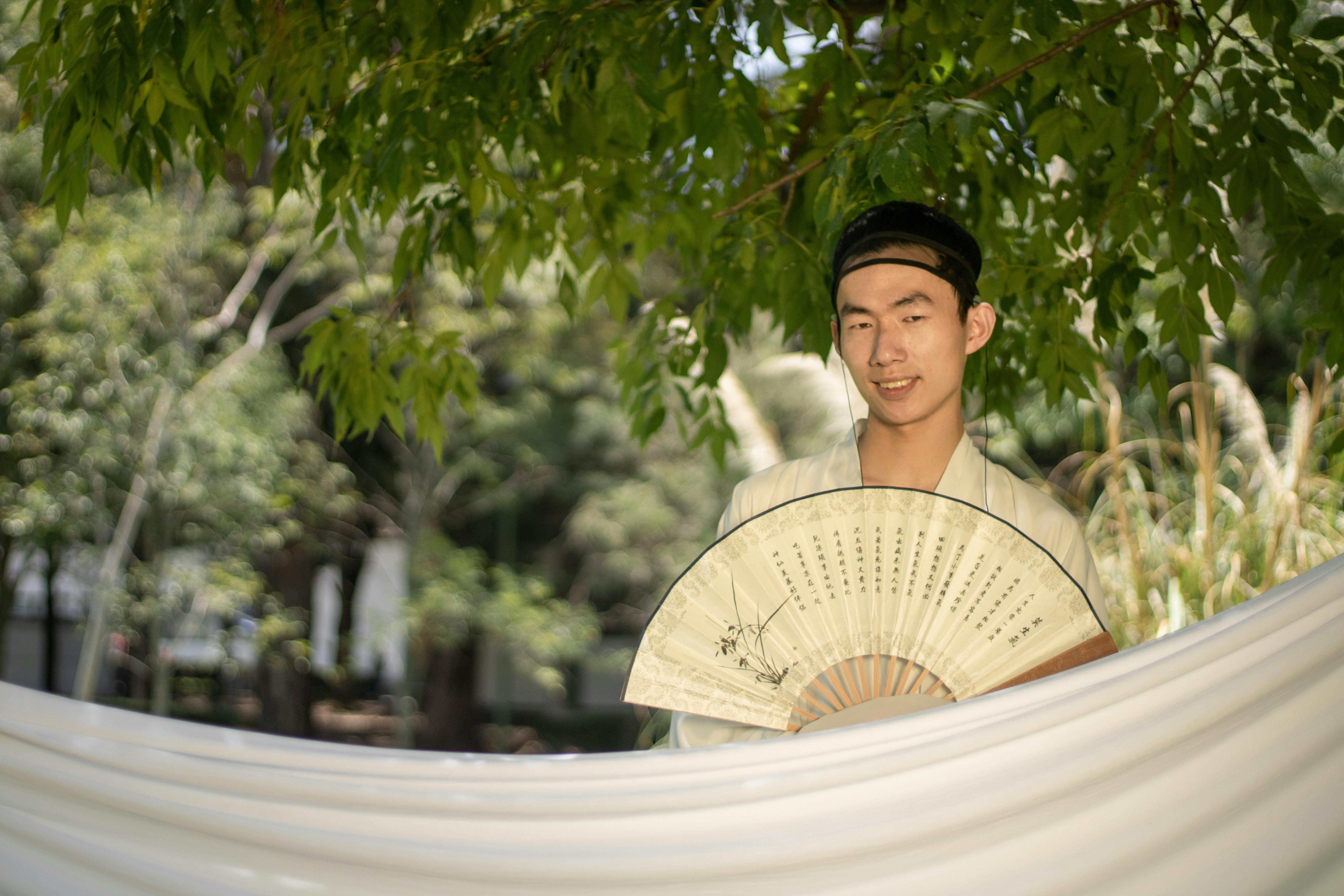 grátis Jovem asiático em traje tradicional com um leque, sob árvores em um jardim sereno. Foto profissional