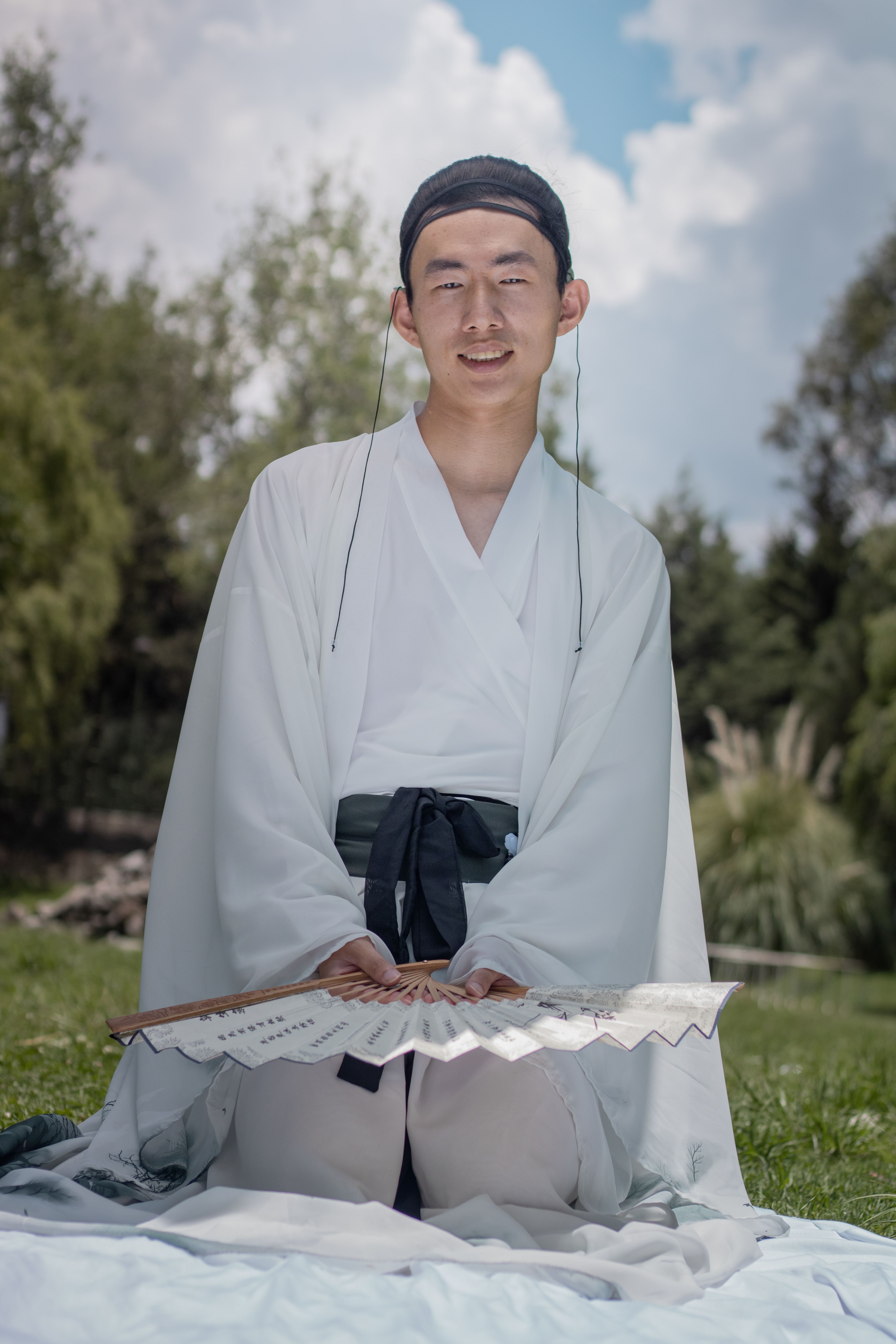 Young man in traditional Asian robe with fan sitting outdoors in nature.