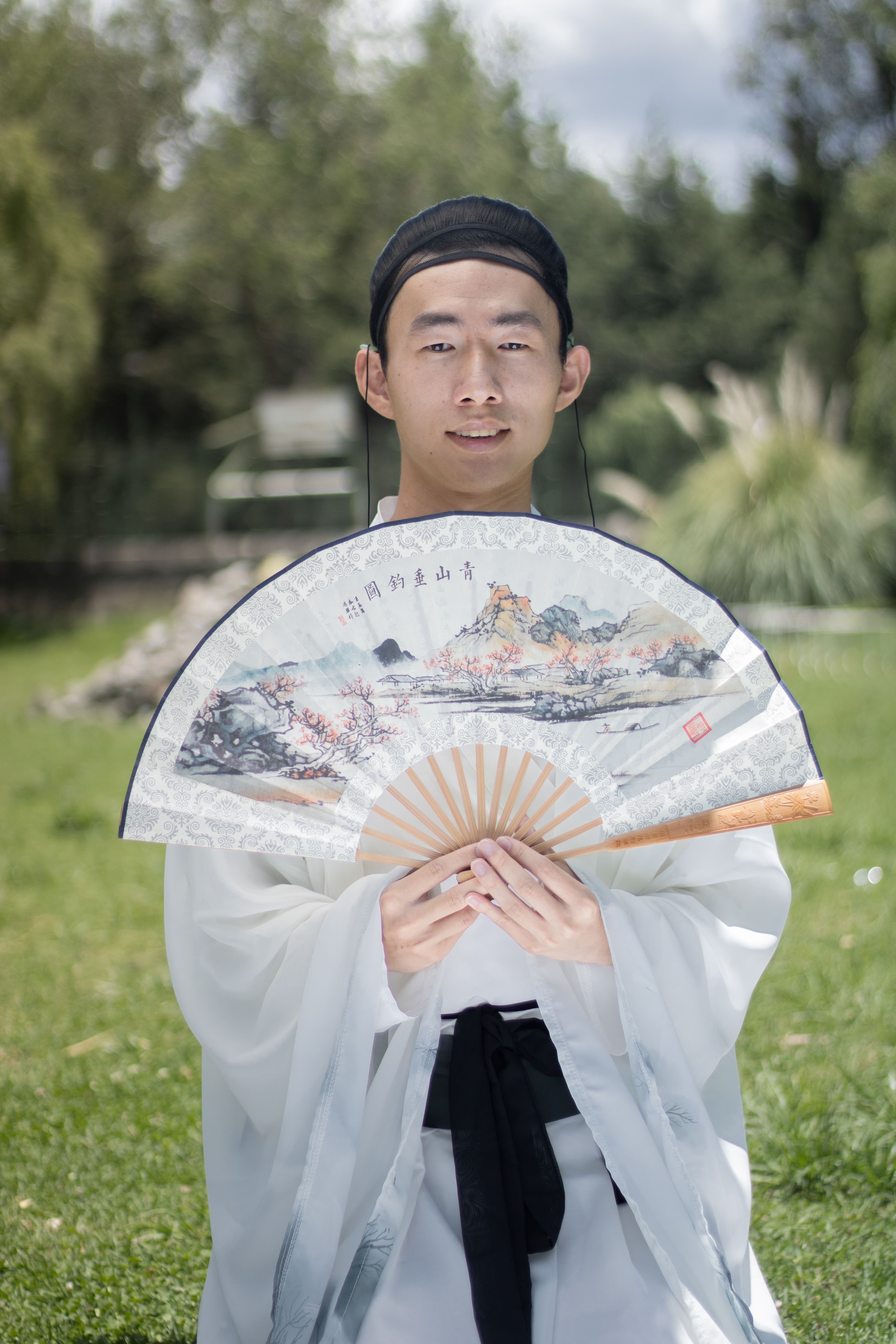 Young man in Hanfu holding a traditional fan, outdoors in nature.