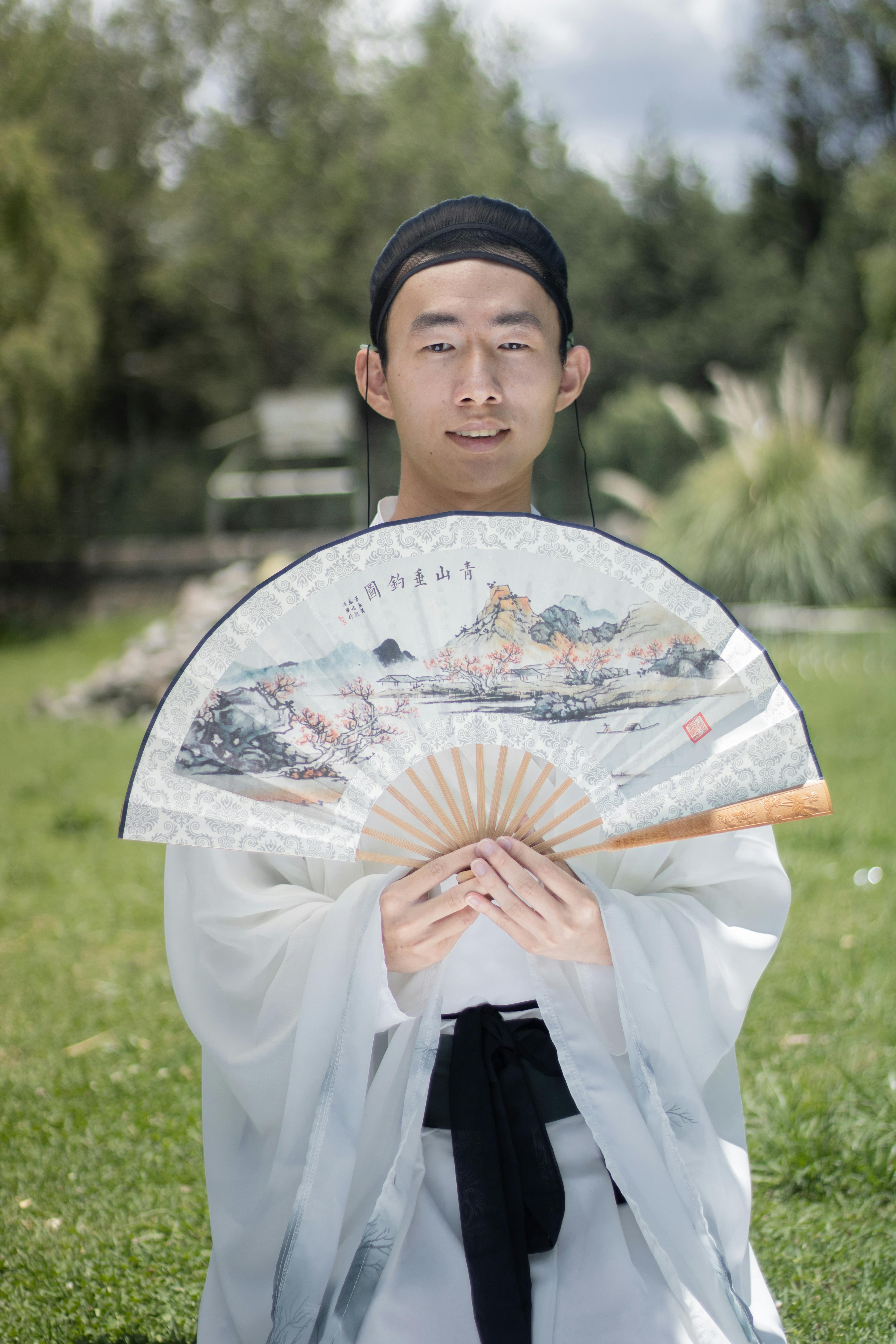 grátis Jovem vestido com hanfu segurando um leque tradicional, ao ar livre em meio à natureza. Foto profissional