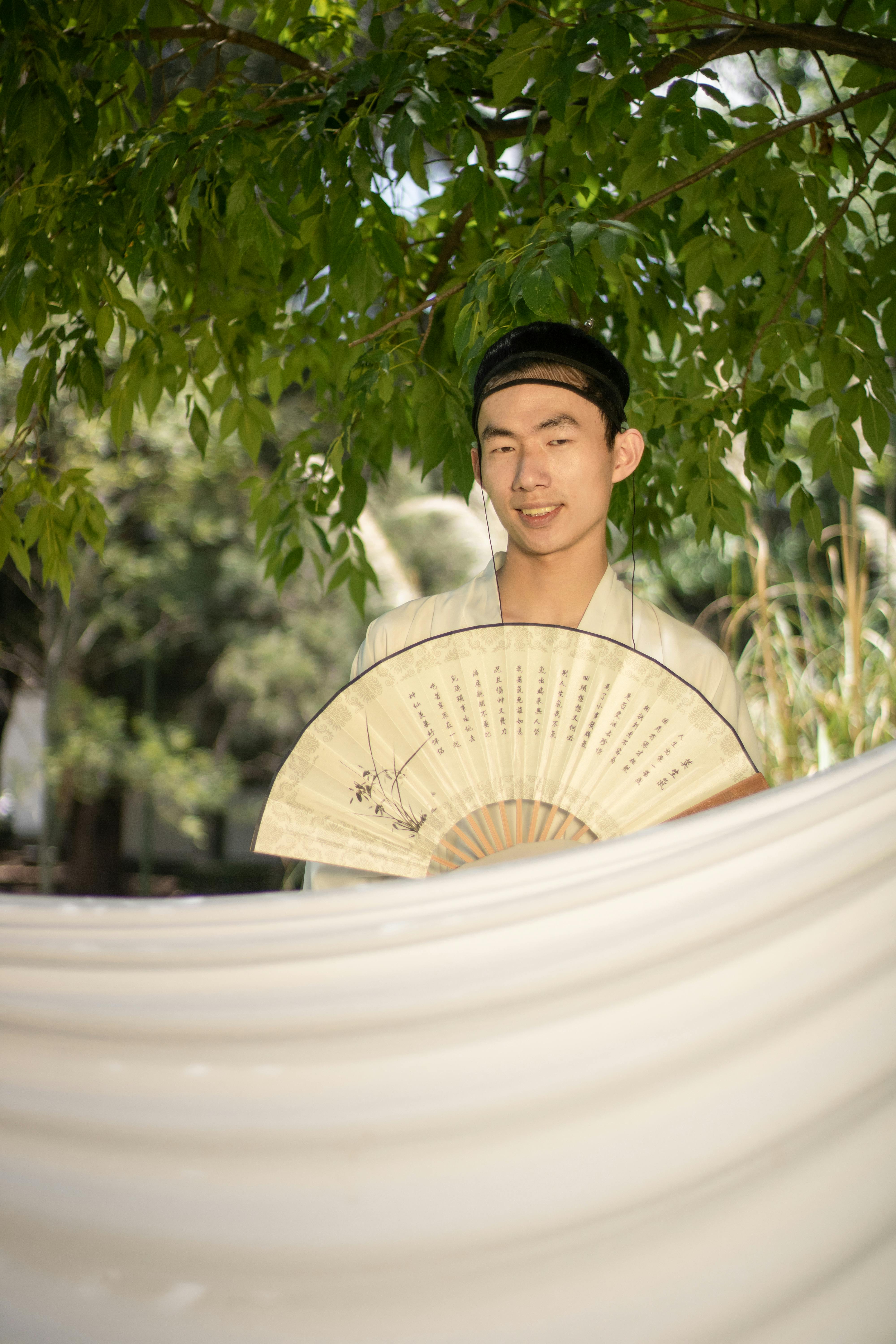 grátis Retrato de um jovem adulto com um leque tradicional chinês sob árvores ao ar livre. Foto profissional