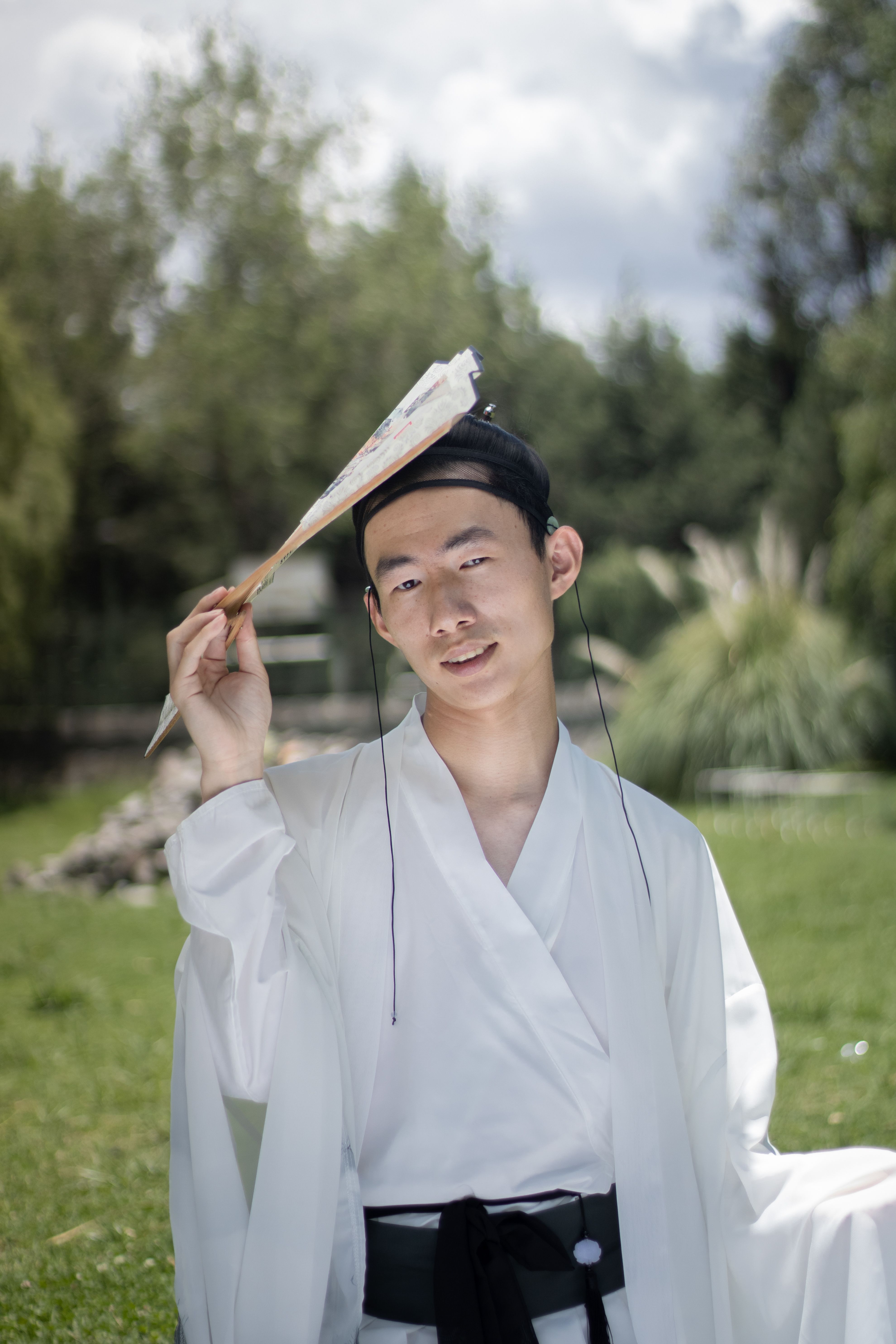 Young man in traditional Asian attire holding a fan outdoors, showcasing culture.